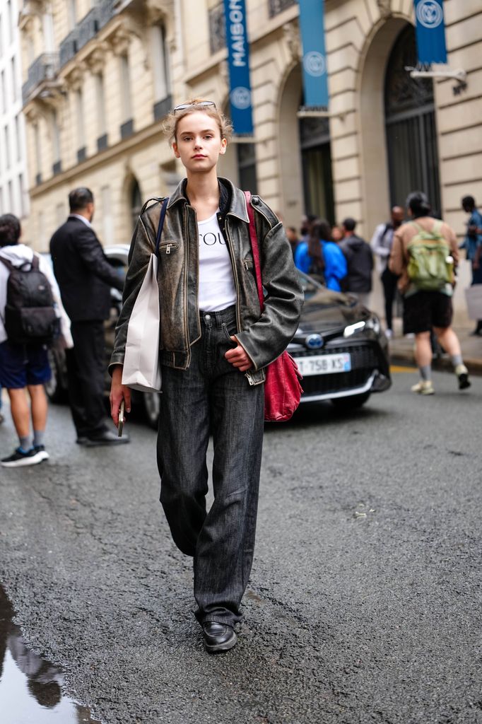 A guest wears distressed black leather jacket with a zip front and zippered pockets is layered over a white T-shirt with visible black lettering from Patou. Dark denim jeans are worn, featuring a straight cut. A red leather shoulder bag with a textured finish is carried on one shoulder. 