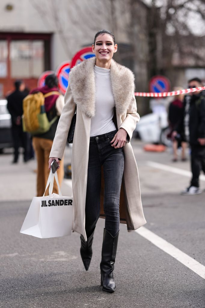 A model wears cream wool coat with light grey faux fur collar, a black denim jean skinny pants, shiny black pointed toe heeled cowboy boots leather shoes, outside Jil Sander.