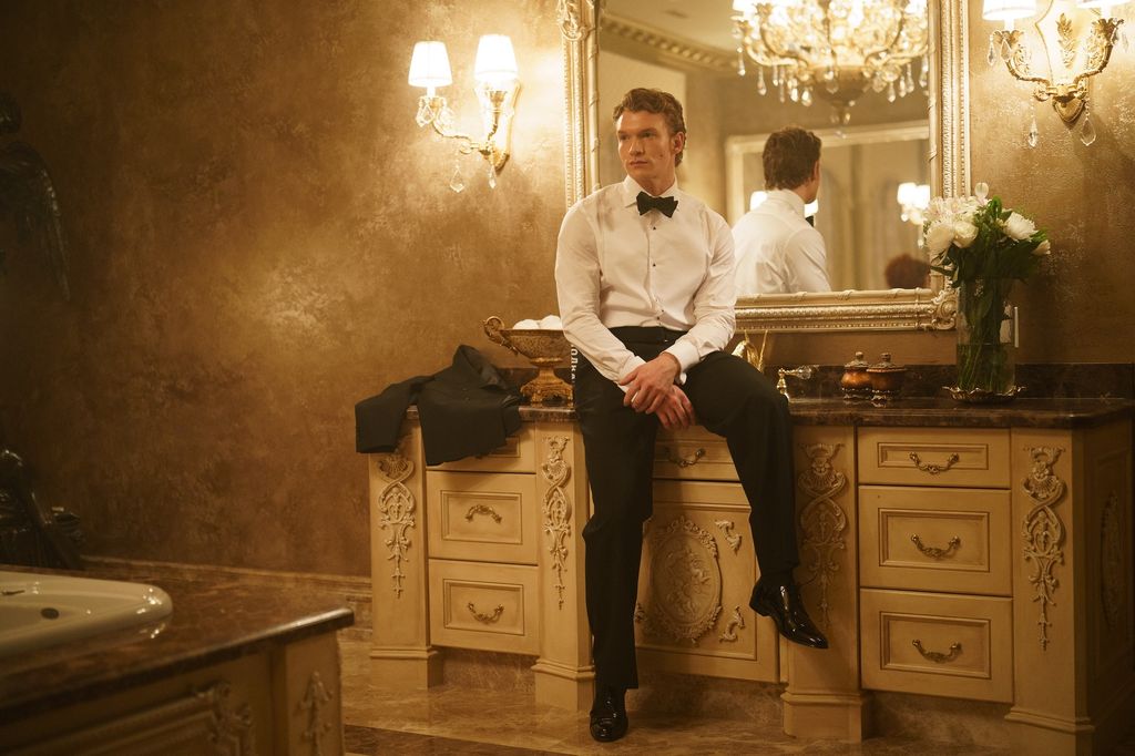 Connor Storrie as Ilya Rozanov sitting on a desk in a tuxedo