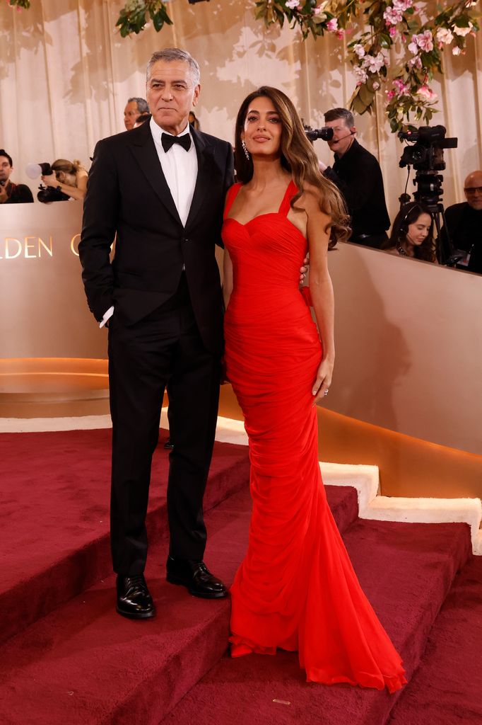 George and Amal Clooney pose on the red carpet - George wears a black blazer, white shirt and black bow tie and Amal wears a long red gown with thin straps