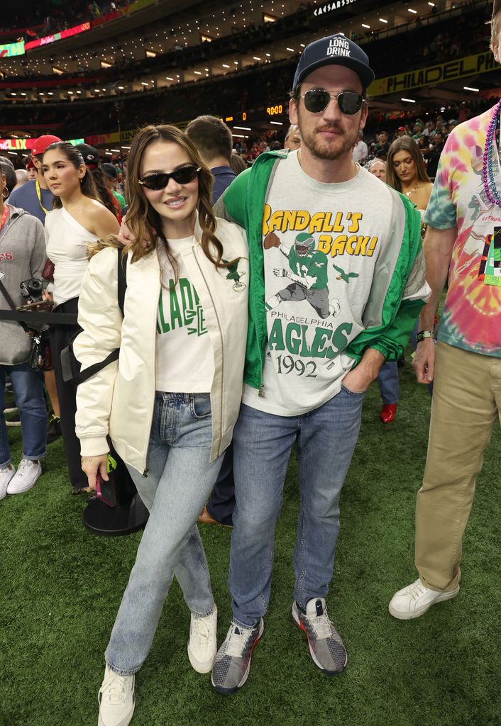 Miles Teller and Keleigh Sperry pose on the Super Bowl field, wearing matching Philadelphia Eagles gear—she in a white bomber jacket and jeans, he in a vintage Eagles sweatshirt and green jacket.