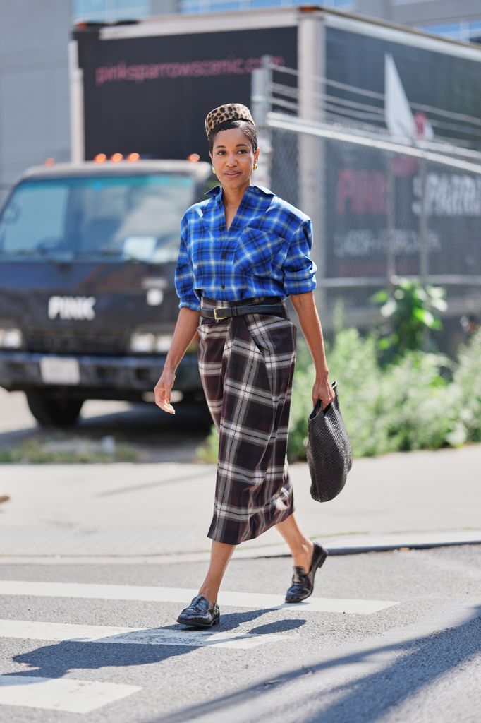A guest wears short natural hair styled under a leopard-print pillbox hat, small gold hoop earrings, a rectangular woven black leather handbag, a blue and black plaid button-up shirt with rolled sleeves tucked into a high-waisted black, gray and white large-checked midi skirt with pleated wrap detailing secured with a black leather belt with a gold rectangular buckle, black leather loafers with a low block heel