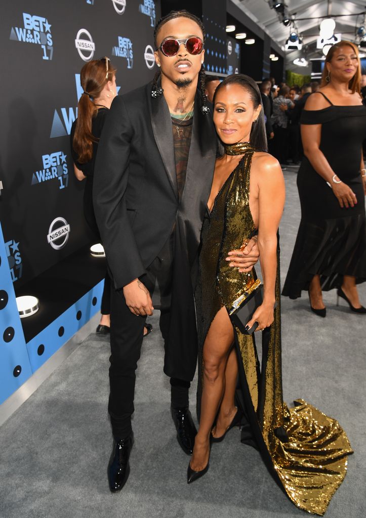 August Alsina wearing a suit (L) and Jada Pinkett Smith wearing a gold halter neck dress