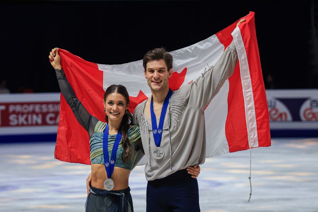 Laurence Fournier Beaudry and Nikolaj Soerensen smiling holding up a Canada flag wearing silver medals on the ice 
