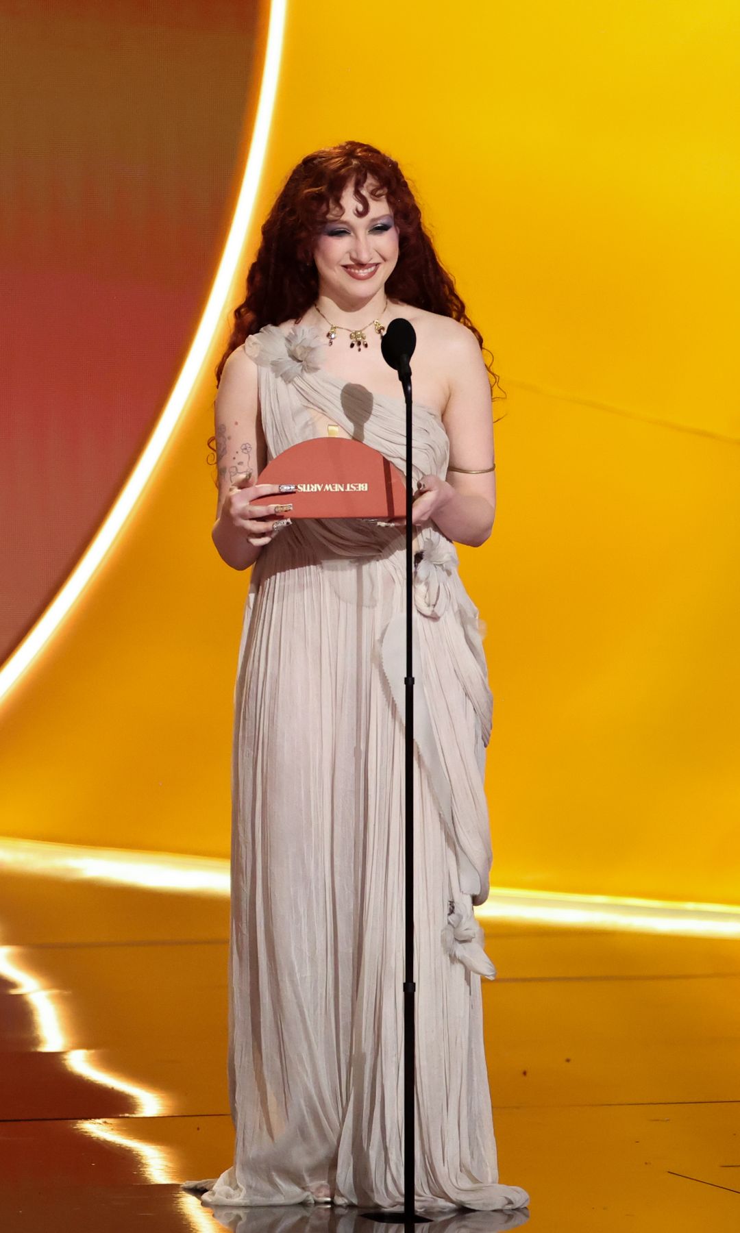 Chappell Roan wears a pale grey sheer draped dress with floral detail as she presents an award on stage