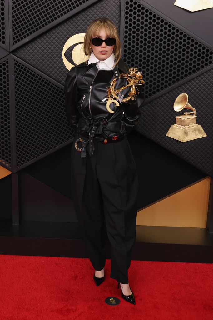 Miley Cyrus wearing a black leather jacket, white shirt, black trousers, black heels and sunglasses and a massive gold brooch