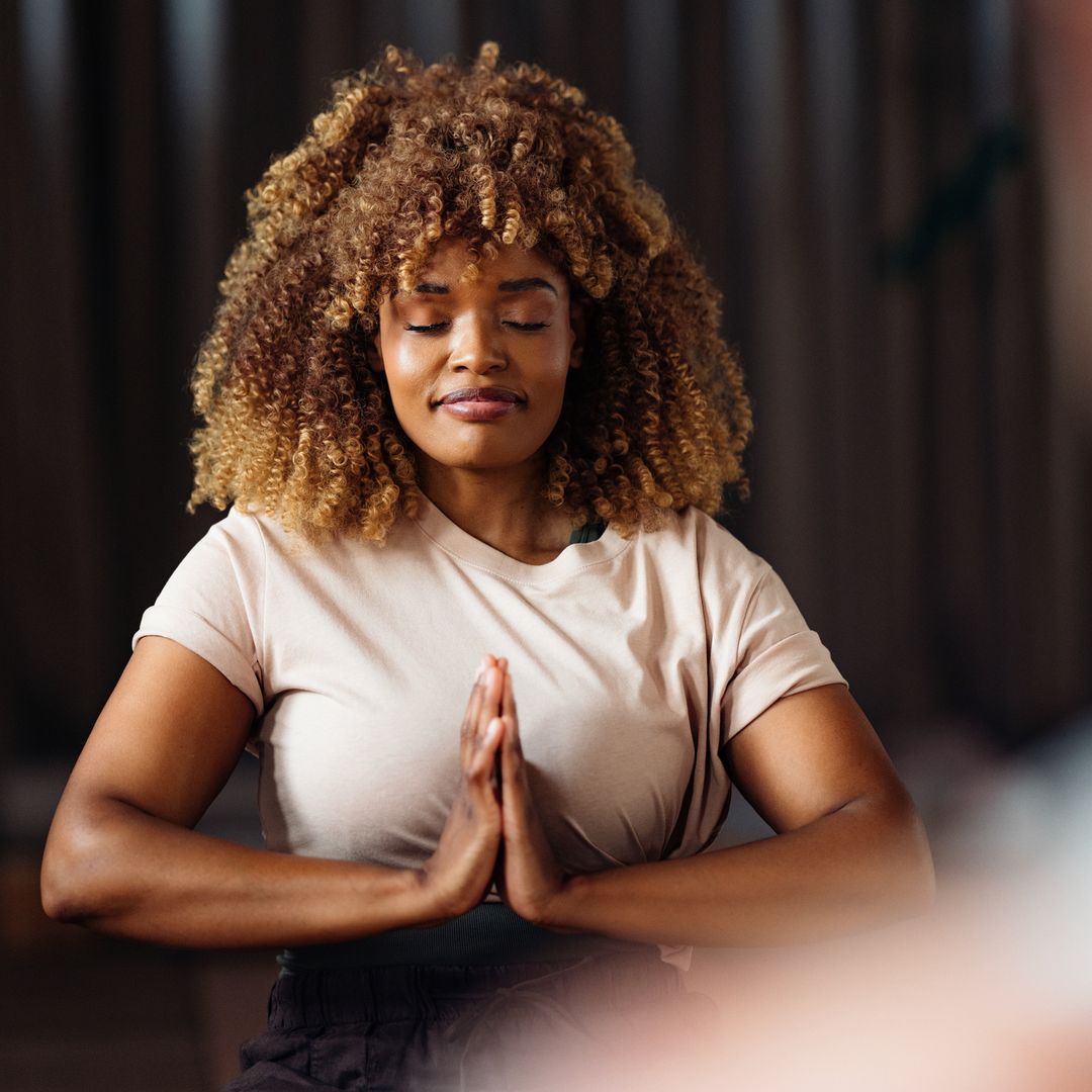 A focused woman sits in a calm yoga class, eyes closed, hands together in a prayer pose, creating a serene moment of mindfulness and balance in a softly lit studio.
