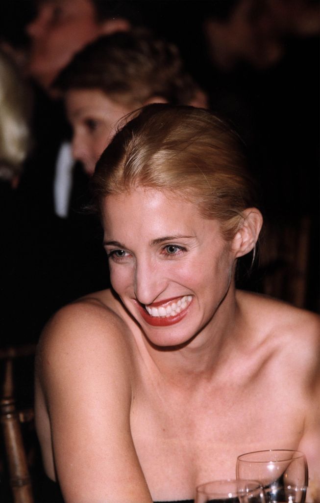 Carolyn Bessette-Kennedy at the Grand Central Station Gala, 1998 in New York City. She laughs at her table, her hair is swept up and she is wearing a black strapless dress