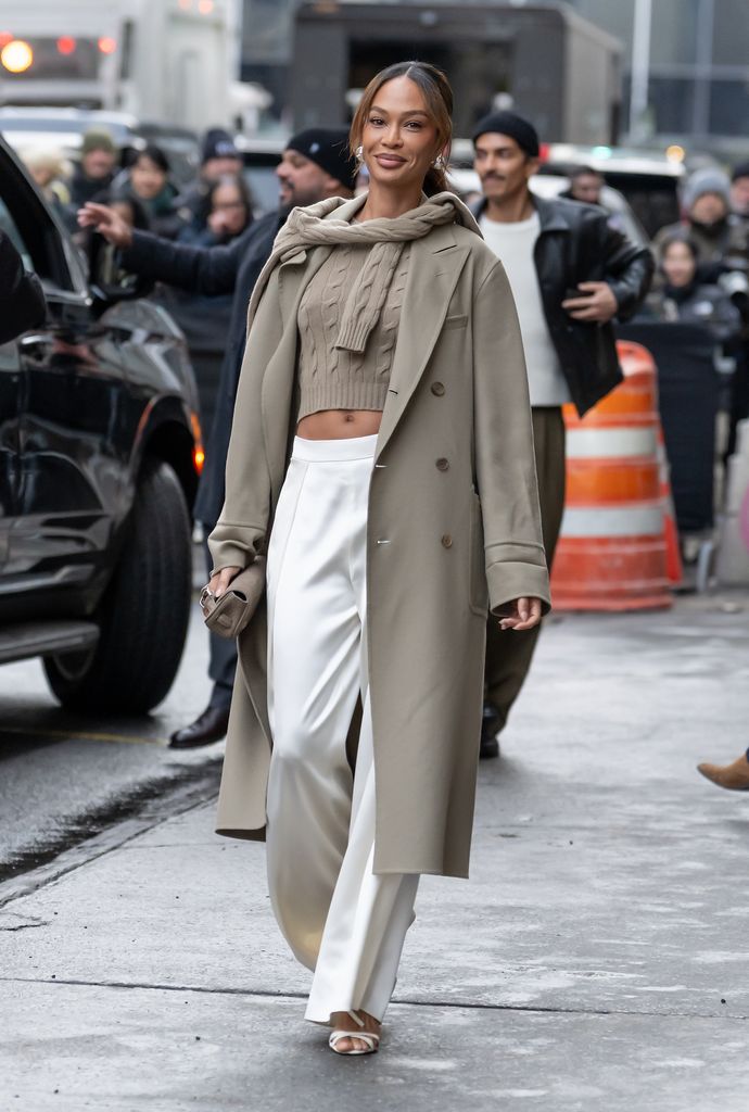 Joan Smalls wears white trousers and a greige matching jacket jumper and second jumper tied around her neck