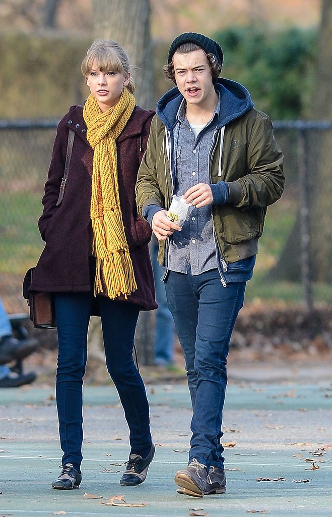 Taylor Swift wears a burgundy coat, a woolly yellow scarf, and jeans as she walks alongside Harry Styles. Harry is dressed in a coat, jeans, and a hat.
