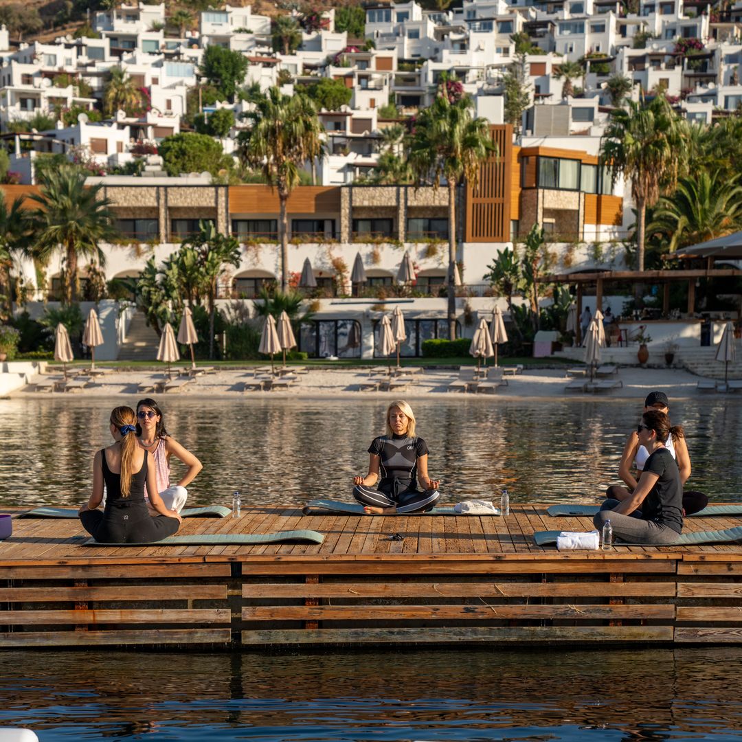 Guests taking part in a morning yoga class on a sea-view deck at Avantgarde Refined Bodrum in Yalıkavak, with the beachfront hotel and hillside homes in the background.
