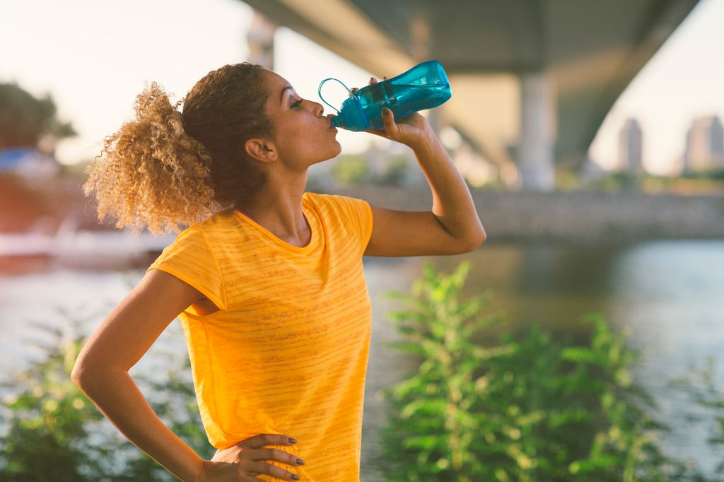 Runner running in the city near river. Standing under the bridge, making break to drink some water. Cityscape in background.