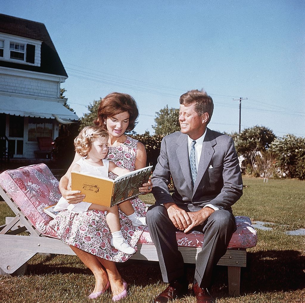Jacqueline Kennedy reads to daughter Caroline who is sat on her lap. Jacqueline and John sit next to one another on a sun lounger.