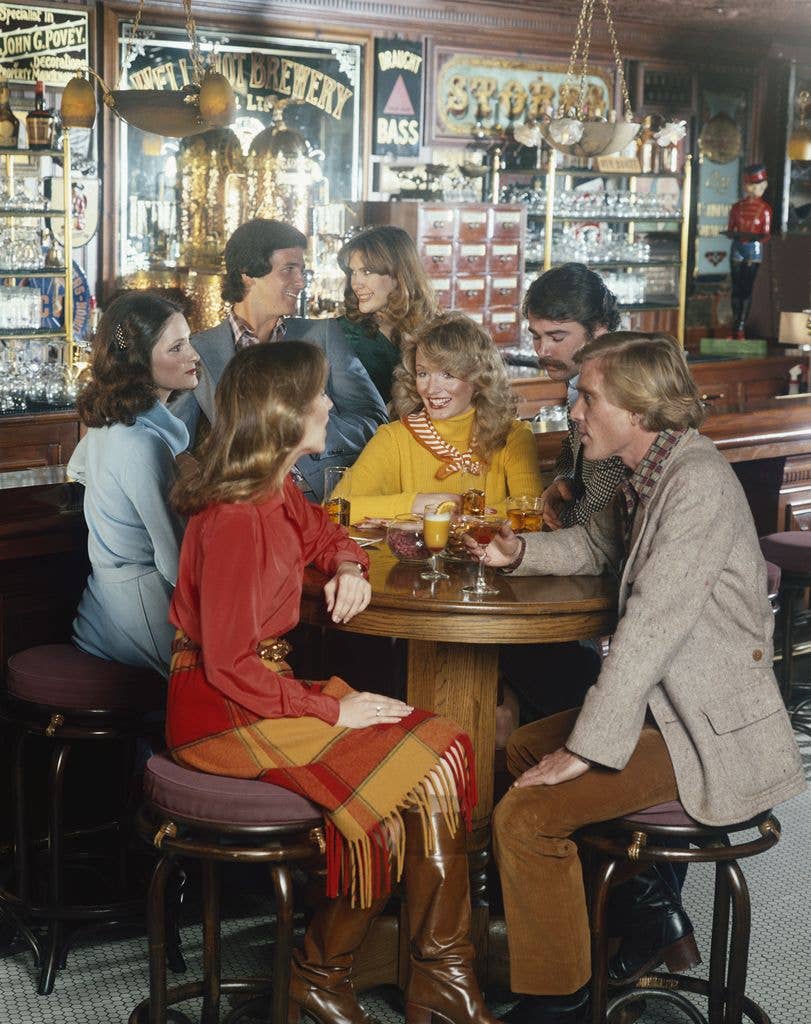 Group of seven people sit around a table in a bar with a mix of drinks on the table.