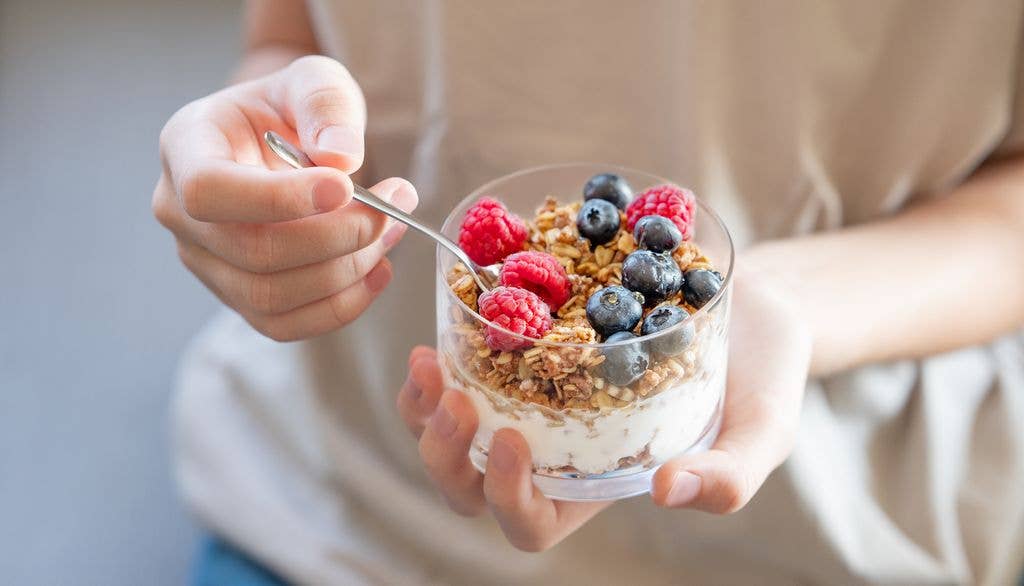 Granola with greek yogurt, raspberries and blueberries in a glass in hands of a woman in a light t-shirt close up. Dairy breakfast concept, oat muesli in the morning light.