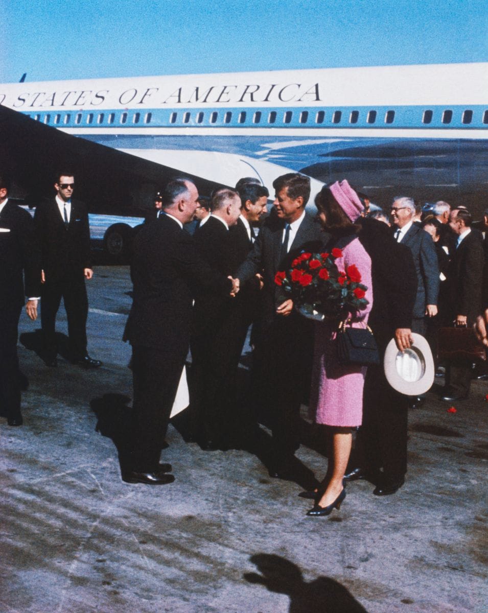 First Lady Jacqueline Kennedy, carrying a bouquet of red roses. The First Lady is wearing a pink suit with a matching hat. Hours later, the President was assassinated.