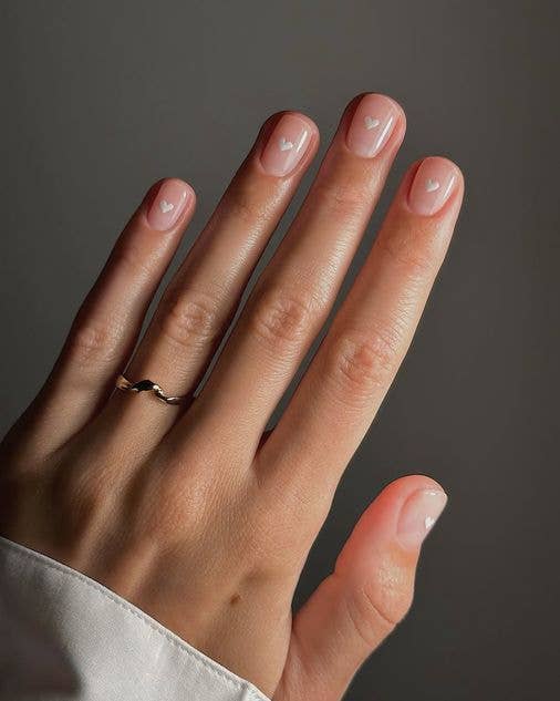 Minimalist nude manicure on short nails decorated with tiny, hand-painted white heart motifs.
