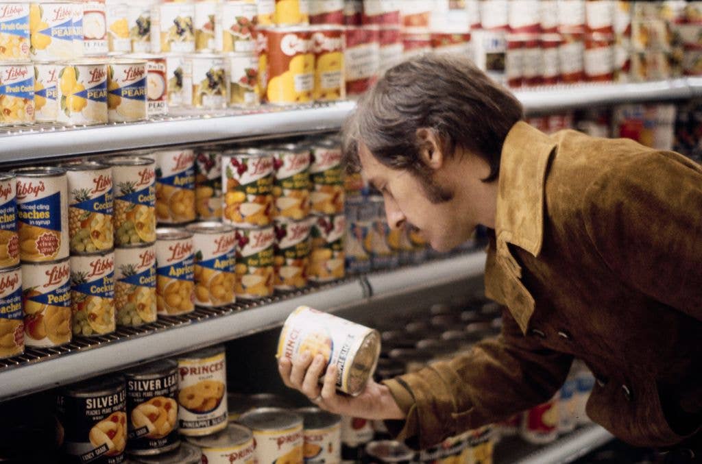 Artist Ed Ruscha shops for produce. He holds a can in his hand and appears to read the label with shelves of canned food on shelves in front of him.