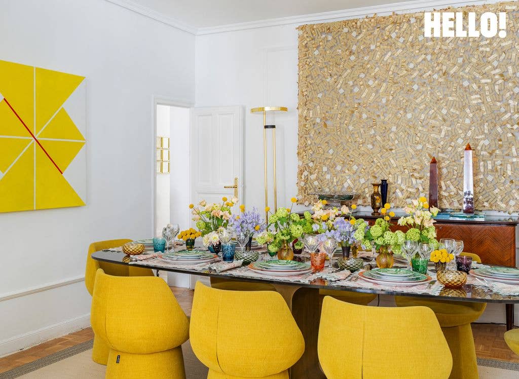 The kitchen in designer Silvia Tcherassi’s Madrid home features bright yellow chairs around a table laid with a dinner set and placemats, complemented by gold and yellow-and-white art pieces on the wall