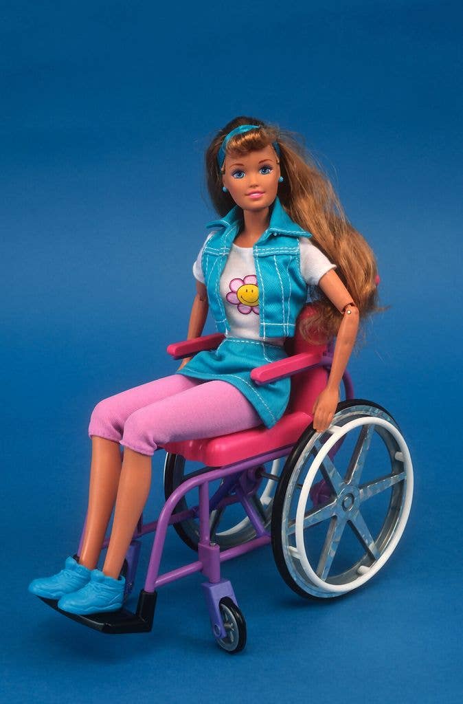Barbie sits in a pink and purple wheelchair wearing a denim skirt and waistcoat with a flower print tee, pink leggings and blue shoes.