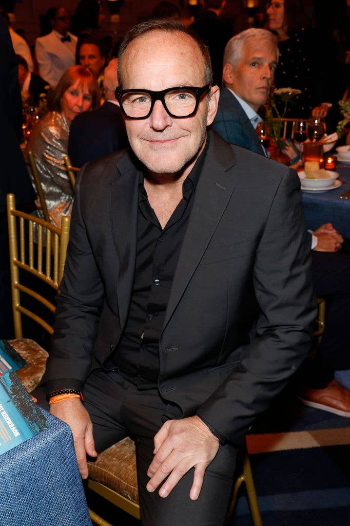 Clark Gregg sat on a chair wearing a black suit and shirt with thick framed black glasses, holding his hands on his thighs.