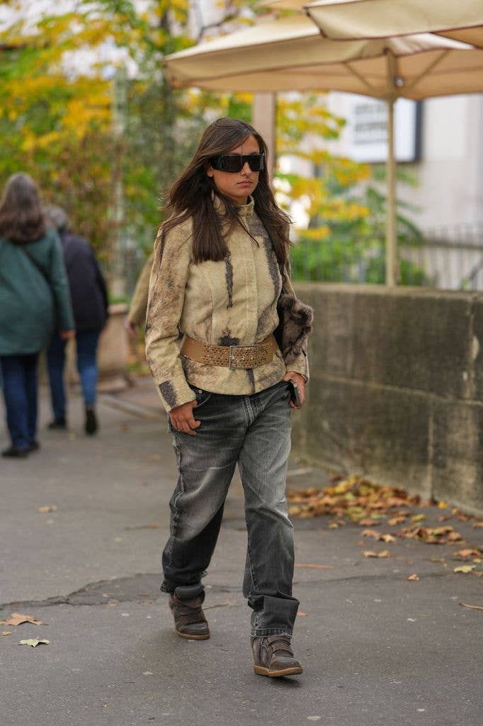 A guest wears long straight dark brown hair with a slight center part, large rectangular black futurist sunglasses, a brown fur clutch bag, a beige shearling jacket with darker brown vertical markings and a high collar, a wide tan leather belt with punched studs and a large rectangular metal buckle, faded grey-black relaxed-fit denim jeans, brown suede wedge sneakers shoes with velcro straps and rubber soles from Isabel Marant, outside Akris, during Paris Fashion Week - Womenswear Spring Summer 2026, on October 05, 2025 in Paris, France (Photo by Edward Berthelot/Getty Images)