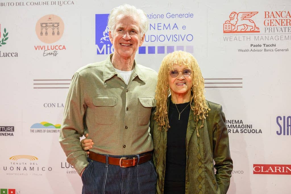 Actor Matthew Modine wears a buttoned up green shirt tucked into blue trousers, held with a brown belt, and a white tshirt. He holds an arm around his wife Caridad who wears a loose green jacket and black dress, with glasses and blonde dreadlocks.