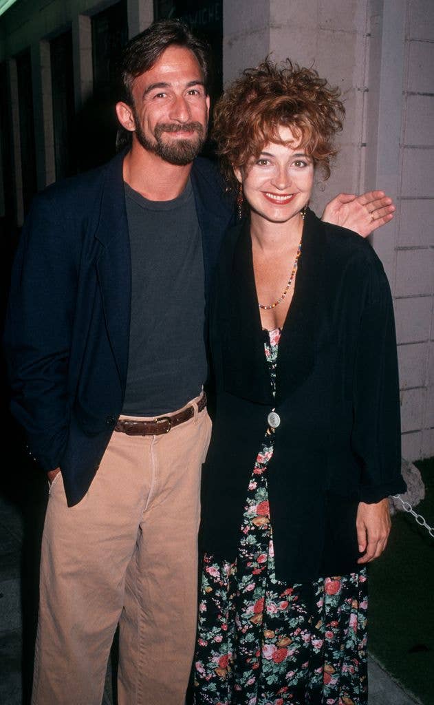 Annie Potts wears a black dress with a colourful ditsy floral print and a loose oversized black suit jacket. She stands with her arm around husband James' waist while he holds a hand out over her shoulder. He wears a black tshirt, black suit jacket and khaki trousers with a brown belt.