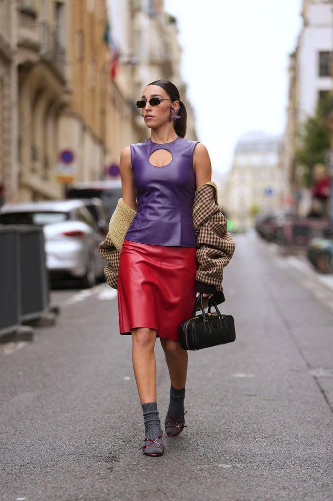 Ginevra Mavilla wears slicked-back dark brown hair in a low bun, sculptural purple earrings, a black leather top-handle bag from Miu Miu with gold hardware, an oversized brown houndstooth wool coat with beige shearling lining, a sleeveless purple leather top with a round keyhole cutout at the chest, a red leather knee-length skirt, gray ribbed socks paired with burgundy leather strappy sandals