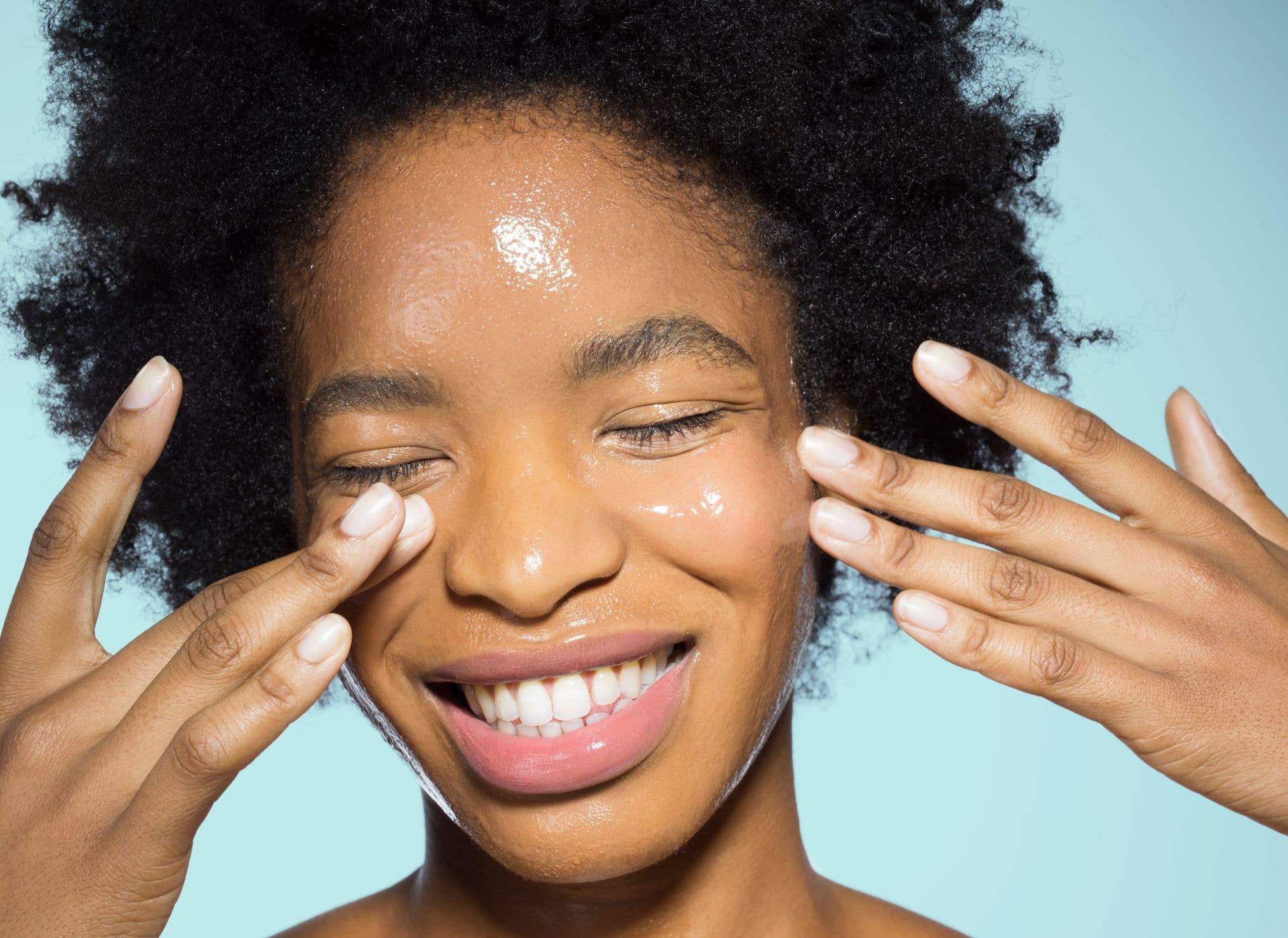 Close-up of young mixed-race woman applying glossy face make-up moisturizer, smiling, with eyes closed with turquoise background