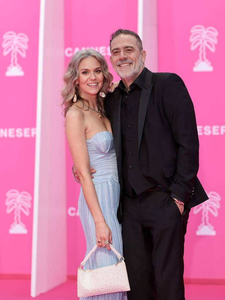 Hilarie Burton and Jeffrey Dean Morgan on the Pink Carpet at Cannes. Jeffrey wears an all black suit and holds an arm around his wife Hilarie who faces into her husband in a light blue pastel dress and holds a white bag, turning her head to face the camera.