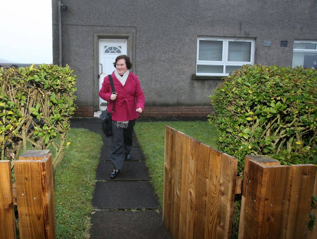 woman in pink jacket walking on path outside grey house towards wooden fence