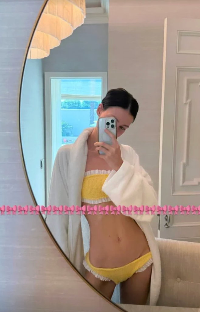 Nicola Peltz posing in the mirror wearing a yellow and white two-piece bikini and a robe 