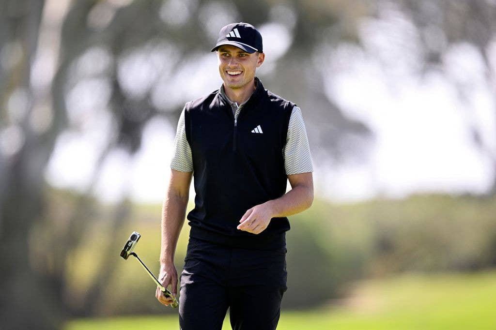 Ludvig Åberg walks across a golf green, with his left hand bent at the elbow and rested at his waist while his right hand carries a golf club horizontally. He smiles and looks to the left of the image, he wears a grey shirt and a black Adidas gilet and matching cap.