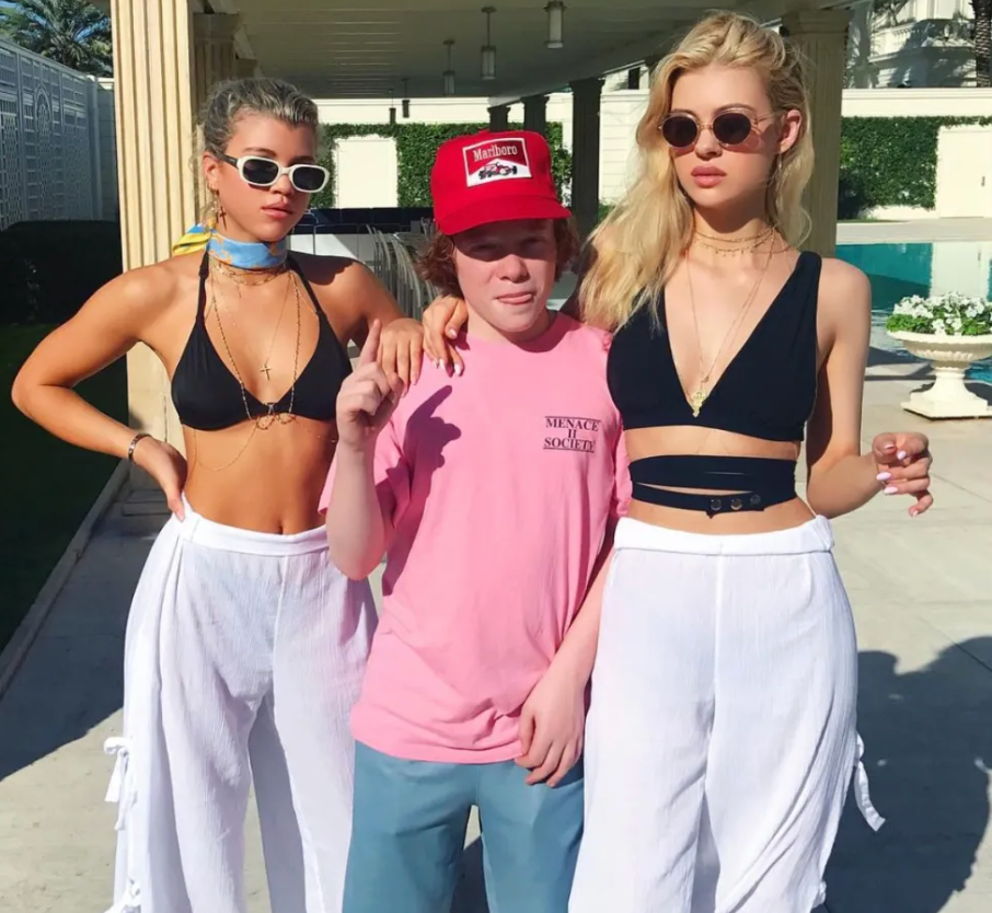 Sofia Richie and Nicola Peltz in black bikinis, white trousers and shades, with a friend in the middle of them