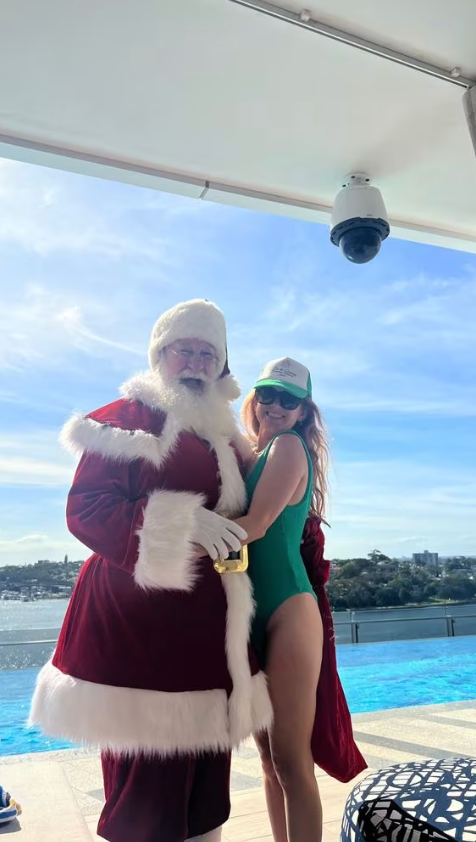 Isla Fisher posing in a green swimsuit by the pool, standing next to Santa Claus
