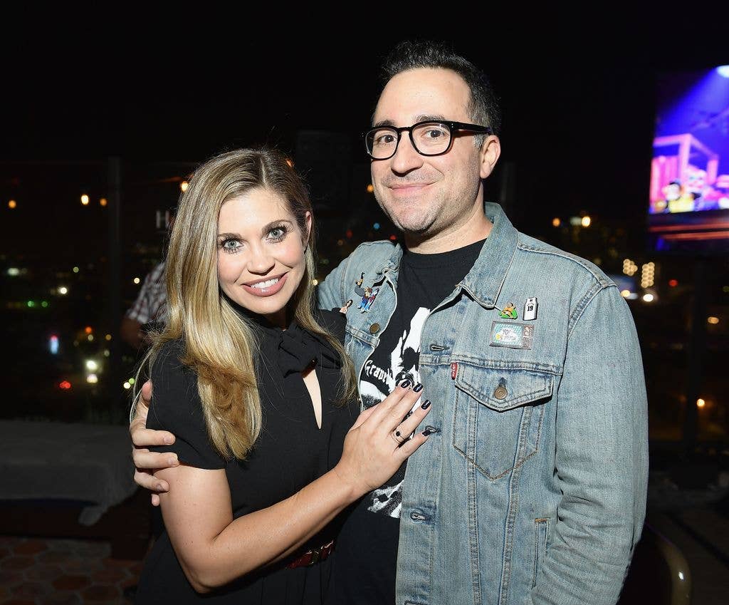 Danielle Fishel holds a hand to husband Jensen Karp's chest while he wraps an arm around her upper back. Danielle wears a black dress and Jensen wears a black printed tshirt with a denim jacket.