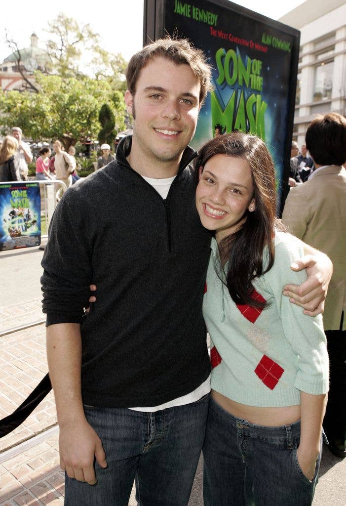 Clay Senechal wears a black quarter zip over a white tshirt and jeans with the sleeves rolled up past his elbows. He holds his arm around Misti Traya who wears a teal jumper with a red and pink diamond pattern and jeans, holding her right arm around Clay and her left hand in her pocket.
