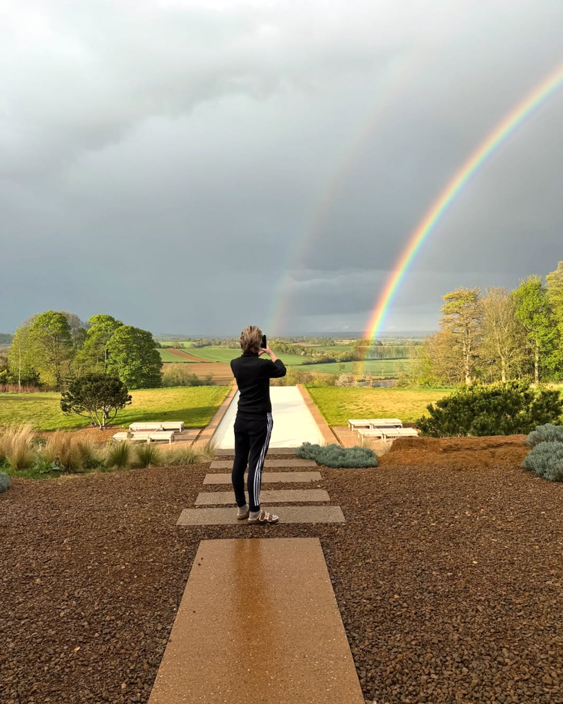 Photo shared by Ellen DeGeneres on Instagram April 2025 of her wife Portia De Rossi taking a photo of a double rainbow over their home in the Cotswolds