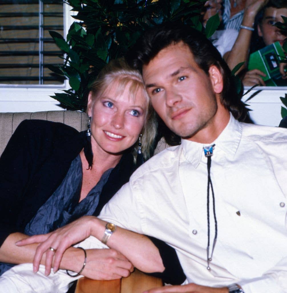 American actor, singer and dancer Patrick Swayze sits in a white shirt gazing to the left of the image and resting his head on wife Lisa Niemi's head who smiles up towards the right hand corner of the image with both hands wrapped around his arm.