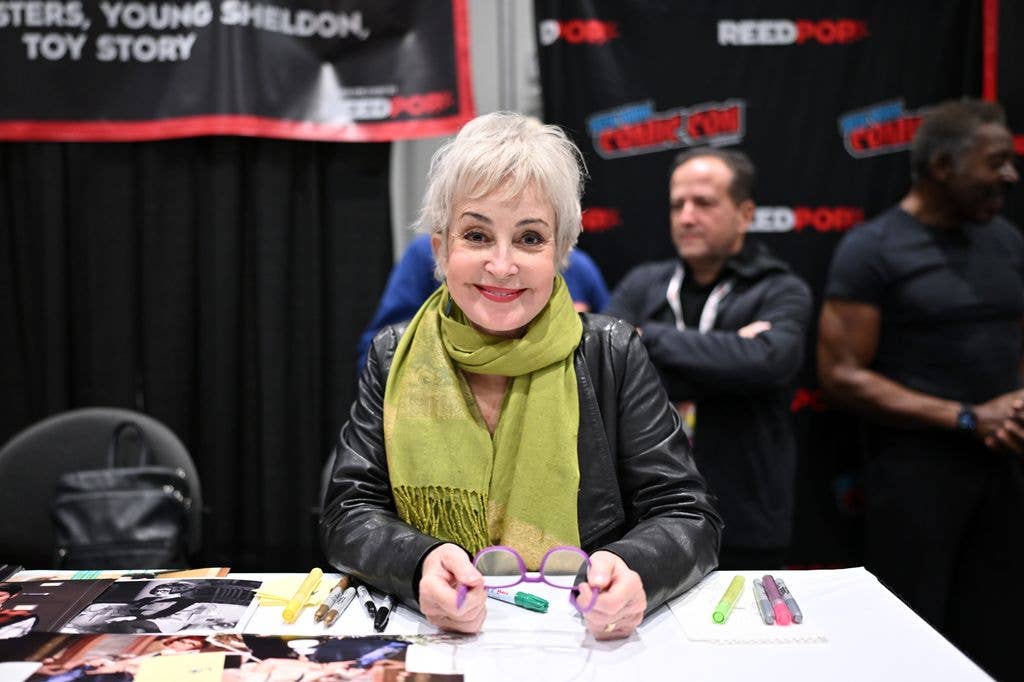 Annie Potts wears a black leather jacket and a green scarf and sits behind a desk at Comic Con holding a pair of purple glasses between her hands.