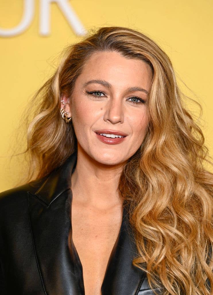 Blake Lively smiles on the red carpet in a black leather jacket dress with voluminous waves and radiant makeup, attending the premiere of A Simple Favour 2.