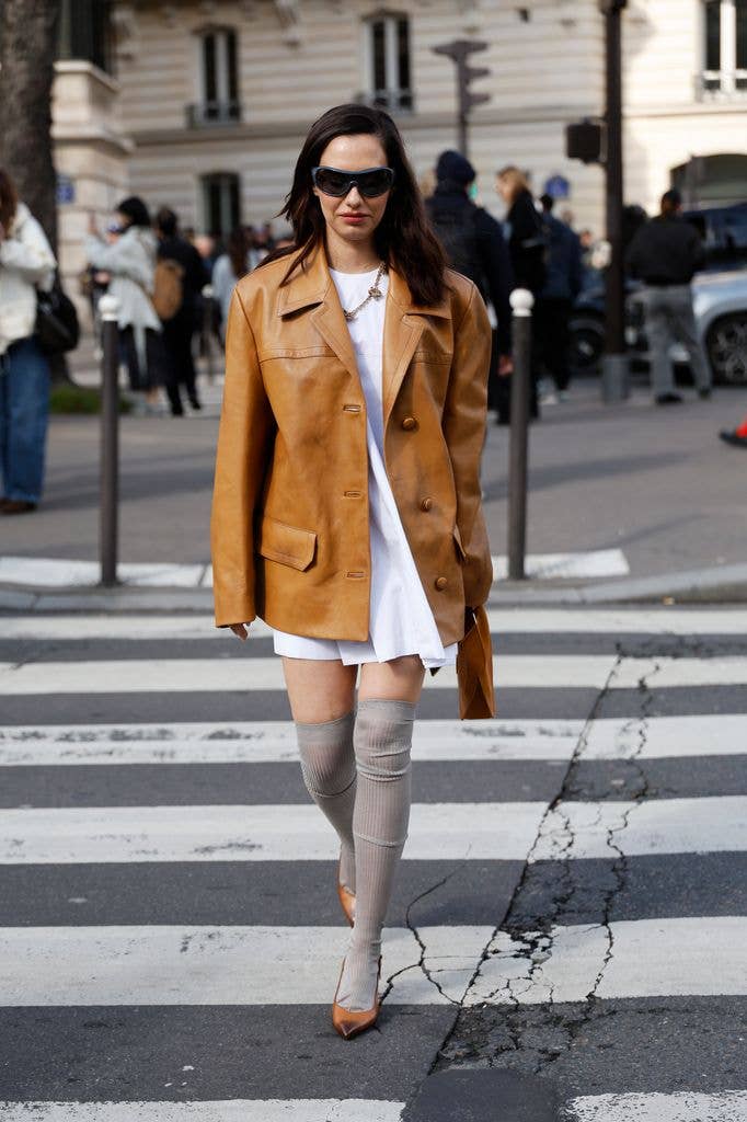 guest wears white mini dress, beige leather blazer, grey ribbed over the knee socks, brown pumps, camel bag, outside Miu Miu, during Day Nine of Paris Fashion Week
