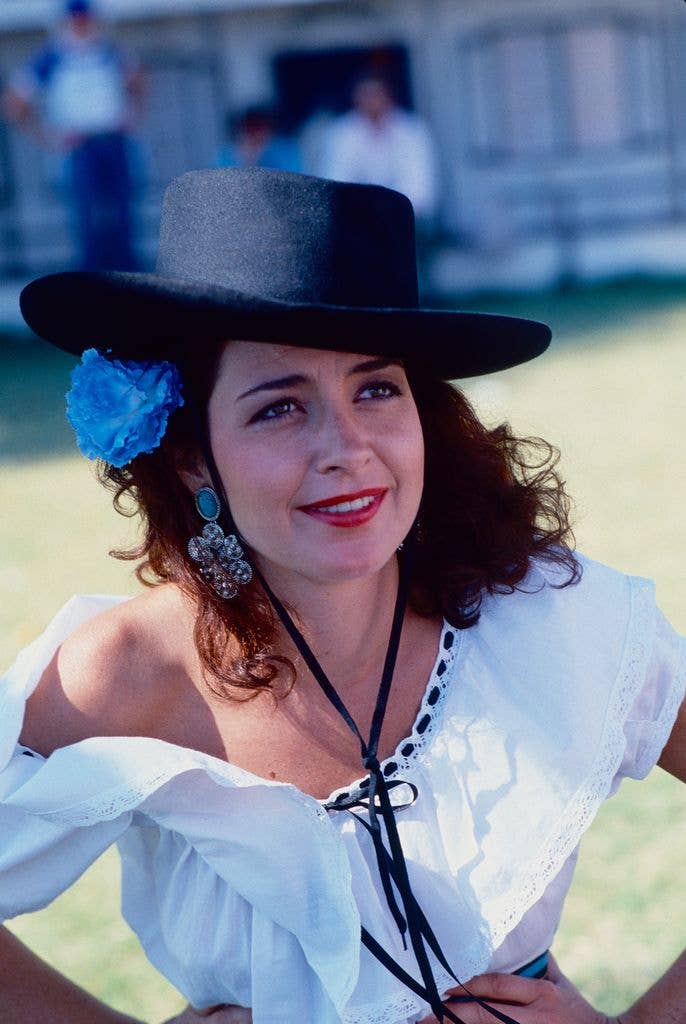 Annie Botts in a white blouse trimmed with black ribbon and one shoulder exposed, wearing a black cowboy hat at an angle and a large blue flower in her hair beneath its brim.