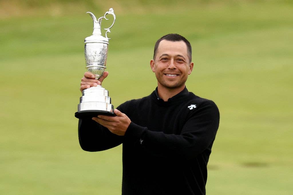 Xander Schauffele holds the Claret Jug trophy aloft with his right hand, supporting the base with his left, and smiles at the camera. He wears a black jacket with a white logo on its left shoulder.