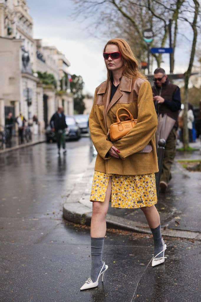  guest wears long red hair, red sunglasses, a brown leather and brown suede oversized jacket, a yellow purple floral print pattern knees dress, a light orange shiny leather handbag from Miu Miu, gray wool ankle socks white leather high heels shoes, outside Miu Miu
