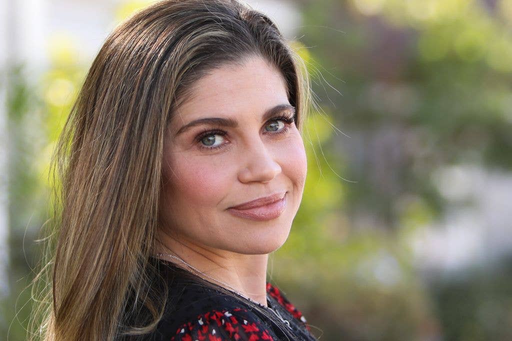 Headshot of Danielle Fishel who looks at the camera with a slight smile, turning her head towards her right shoulder, backgrounded by blurred trees.