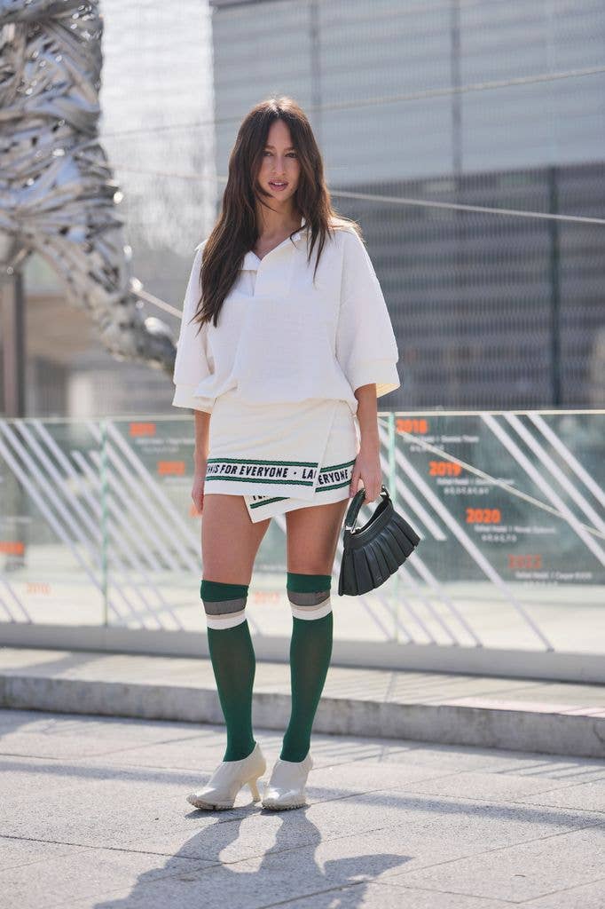 Isabelle Allain wears dark brown hair, a white cotton loose fit polo shirt from Lacoste, a white cotton wrap mini skirt with green and black printed text trim from Lacoste, a dark green leather pleated bag from Lacoste, green knees socks