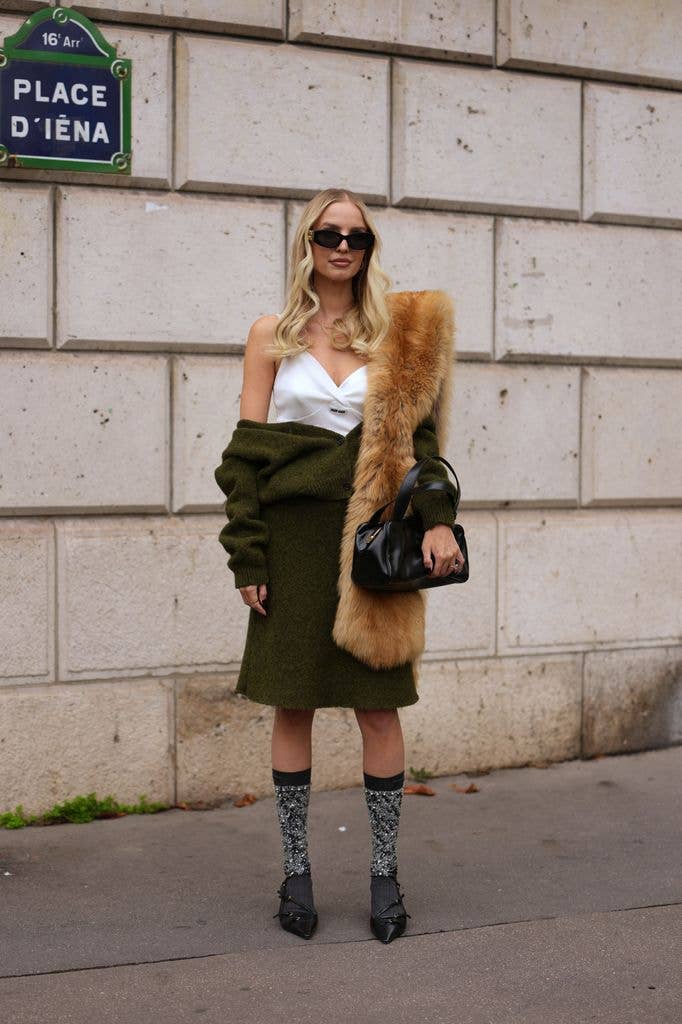 Leonie Hanne wears long blonde hair in loose waves with a center part, black rectangular sunglasses, a gold necklace and gold rings, a black leather shoulder bag from Miu Miu with top handles, a tan fur stole, an olive green wool knit cardigan worn off the shoulders, a white V-neck textured top, an olive green wool knit knee-length skirt, grey and silver sequin knee socks with black top bands, black leather pointed-toe shoes with a small heel, outside Miu Miu