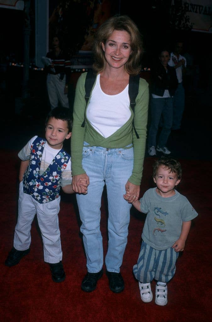 Actress Annie Potts holds the hands of sons Doc Hayman and Harry Hayman. She wears a white tshirt with a green cardigan, a black backpack and jeans. Son Doc wears light jeans, holds his right hand in his pocket and wears a blue patterned waistcoat over a white tshirt. Son Harry stands straight on to the camera in striped teal trousers and a matching green tshirt with white trainers.